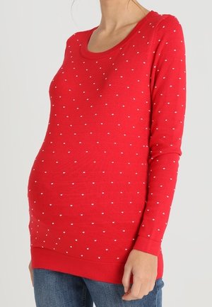 Personne enceinte portant un pull rouge à manches longues avec de petits pois blancs, assorti à un jean bleu, debout devant un fond uni.