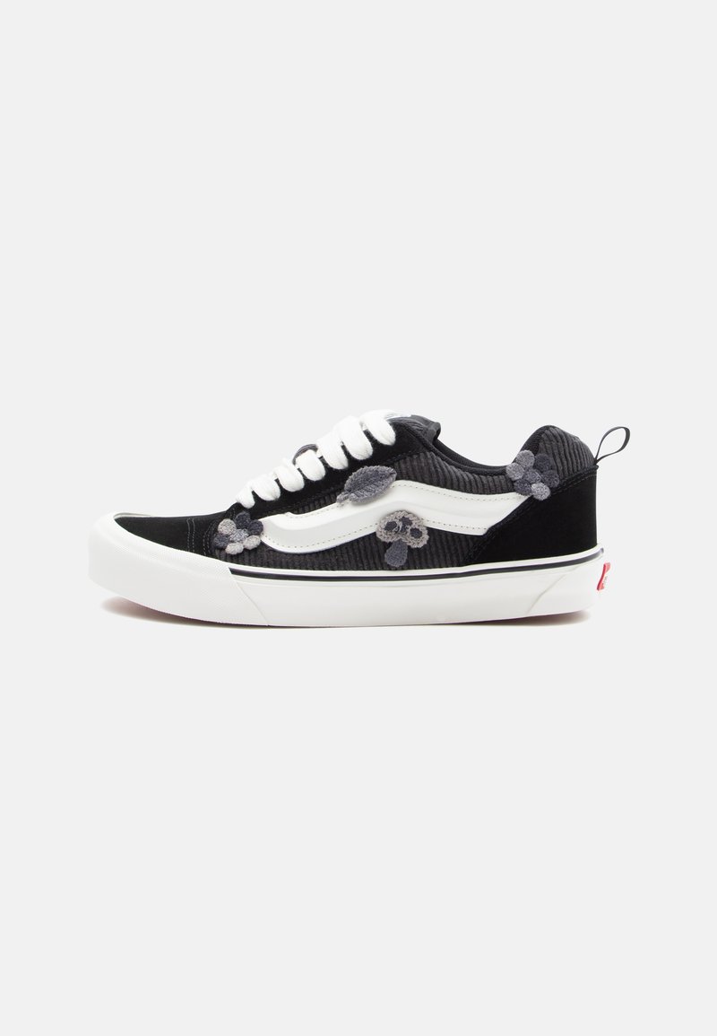 Black corduroy sneaker with white leather accents, featuring floral appliqués in shades of gray and a white rubber sole. Classic lace-up design.