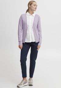 Gilet violet clair, texture côtelée, devant boutonné, porté sur une chemise blanche. Associé à un pantalon navy et des baskets grises. Design simple.