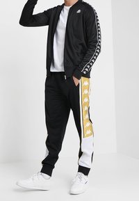 Black zip-up track jacket with white logo, paired with black joggers featuring gold and white side stripes. White sneakers complete the look.