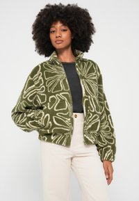 Chaqueta de forro polar verde y aterciopelada con patrones florales blancos, cremallera frontal y cuello alto. Llevada sobre una camiseta negra, combinada con pantalones claros.