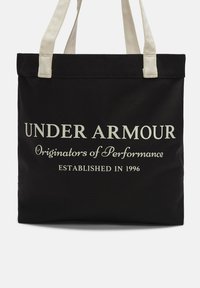 Borsa tote nera in tela con manici in tessuto beige. Presenta un grande testo bianco che recita "UNDER ARMOUR," "Originatori delle Performance," e "FONDATO NEL 1996."