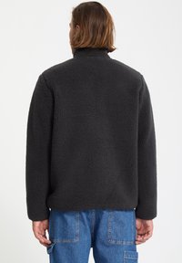 Veste en polaire noire avec un col haut, un extérieur texturé et une coupe décontractée ; elle présente un ourlet lâche et des manches longues.