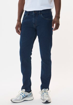 Jeans de denim azul oscuro con corte recto, diseño de cinco bolsillos y costuras en contraste. Llevados con zapatillas deportivas grises.