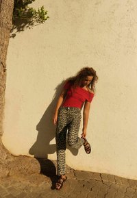 Sarkans off-shoulder tops, kas apvienots ar leopardu raksta biksēm un melniem sandaliem. Fona teksturēta siena un koka stumbrs. Saules apgaismota aina.