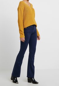 Musztardowo-żółty sweter dzianinowy o fakturze z dekoltem w serek, zestawiony z granatowymi jeansami dzwonami. Modelka nosi czarne botki na obcasie.