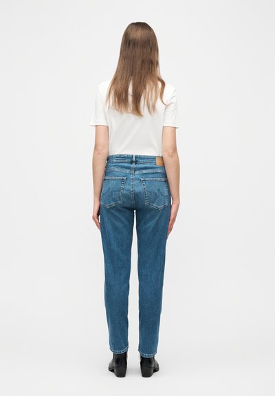 Γυναίκα που στέκεται με την πλάτη γυρισμένη, φοράει λευκό μπλουζάκι με κοντά μανίκια, τζιν ψηλόμεσο μπλε και μαύρα μποτάκια αστραγάλου σε απλό φόντο.