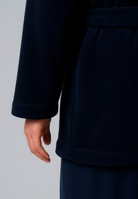 Mano sinistra rilassata al fianco, indossa una giacca a maniche lunghe di un blu navy scuro con cuciture visibili ai polsini e pantaloni abbinati di colore navy.