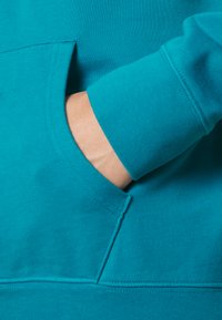 Hand van een persoon gedeeltelijk binnen de voorzak van een turquoise sweatshirt met zichtbare stiksels en geribde manchetten.