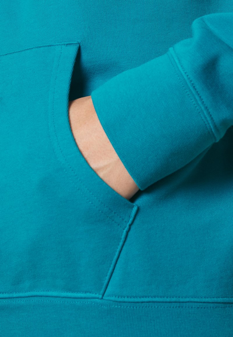La mano de una persona parcialmente dentro del bolsillo frontal de un sudadera turquesa con costuras visibles y puños acanalados.