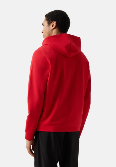 Homme vu de dos portant un sweat à capuche rouge uni et un pantalon noir sur un fond clair uni.