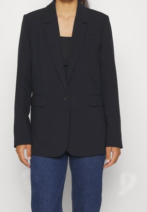 Personne portant un blazer noir à un bouton sur un top noir et un jean bleu, debout devant un fond blanc uni.