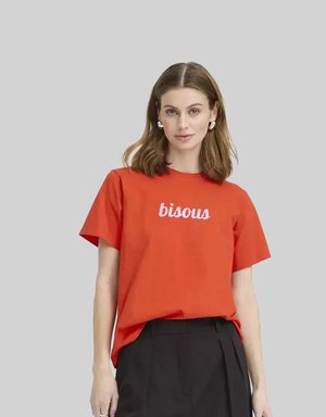 Mujer con cabello castaño hasta los hombros, que lleva pendientes de aro plateados, una camiseta roja con el texto "bisous" y pantalones negros de cintura alta, de pie frente a un fondo gris.