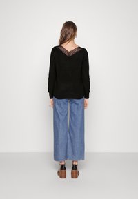 Femme de dos portant un pull noir avec un dos en dentelle en forme de V, un jean large bleu, et des sandales à talons noirs sur fond blanc.