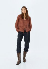 Blusa marrone a trama con orlo arricciato, chiusura con bottoni e maniche lunghe, abbinata a jeans di denim blu scuro e stivali da cowboy ricamati marroni.