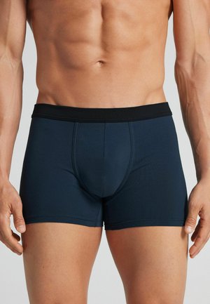 Torso masculin et parties supérieures des jambes portant un boxer bleu foncé avec une ceinture noire sur un fond uni.