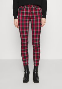 Pantalons slim-fit à carreaux rouges et noirs portés avec un haut noir à manches longues et des bottines noires, montrés de la taille aux pieds sur un fond neutre.