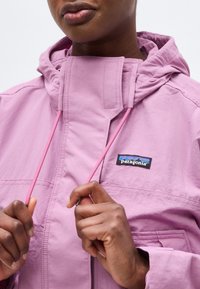 Personne portant une veste Patagonia violet clair, tenant les cordons roses près du cou avec les deux mains, montrant partiellement le visage et le haut du torse.