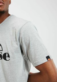 Camiseta de manga corta gris hecha de mezcla de algodón. Presenta un logo impreso en negro y un pequeño parche de logo a color en la manga. Diseño de cuello en V.