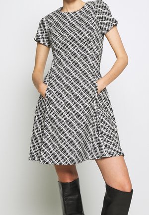 Femme portant une robe à manches courtes à motif noir et blanc avec des poches et des bottes noires montantes, debout devant un fond uni.
