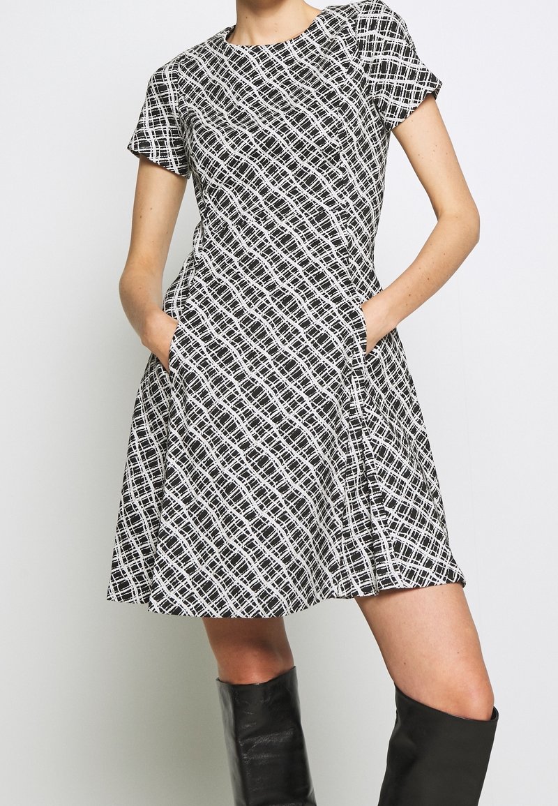 Femme portant une robe à manches courtes à motif noir et blanc avec des poches et des bottes noires montantes, debout devant un fond uni.