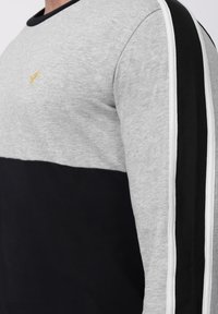 Siva i crna majica s dugim rukavima i okruglim izrezom, s horizontalnim dizajnom blokova boja i malim žutim logotipom na prsima.