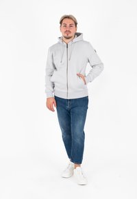 Lichtgrijze hoodie met rits, voorzien van trekkoorden, kangoeroezakken en ribbelmanchetten, gecombineerd met donkerblauwe jeans en witte sneakers.