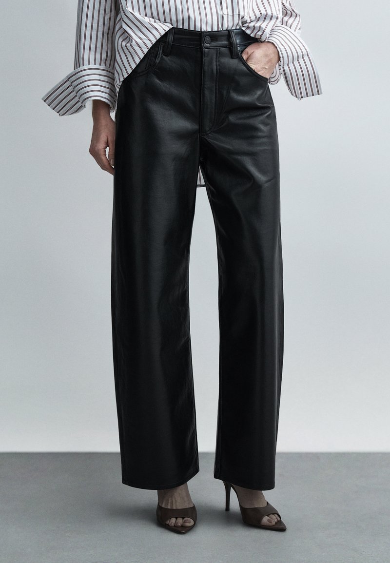 Mustad nahast laialotste püksid, millel on sile tekstuur ja kõrge vöökoht, ees taskud ja nööpidega kinnis, kantud avatud varbaga kontsakingadega.