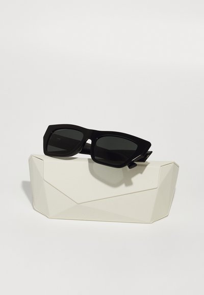 Occhiali da sole neri con montatura rettangolare oversize e lenti scure, appoggiati su un astuccio rigido bianco e angolare.
