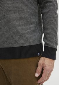 Suéter a rayas en gris y blanco, textura de punto, puños y dobladillo ribeteados en negro, con una etiqueta de la marca en la parte inferior izquierda.