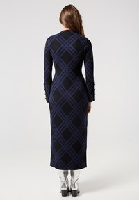 Lungo abito nero a coste con motivo geometrico blu e maniche lunghe, con dettagli a bottoni sui polsini delle maniche. Stivali argento fino al ginocchio.