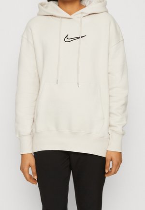 Osoba nosząca kremową bluzę Nike z kapturem i kieszenią z przodu oraz czarne spodnie, stojąca na jednolitym tle.
