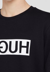 Schwarzes Baumwoll-T-Shirt mit einem weißen rechteckigen Logo, auf dem der Text "HUGO" in Fettdruck steht. Kurze Ärmel und runder Halsausschnitt. Weiche Textur.