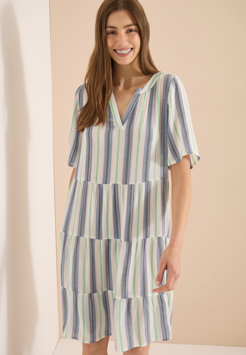 Femme souriante portant une robe ample à manches courtes avec des rayures verticales bleu, vert et blanc, debout devant un fond beige.