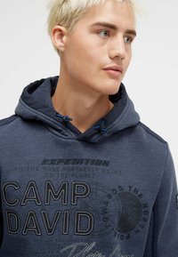 Navy blue hoodie with a drawstring hood, featuring "CAMP DAVID" printed in large letters and expedition-themed graphics on the front.