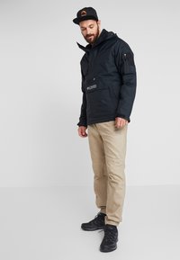 Zwarte pulloverjack met capuchon, voorzien van een voorzak, verstelbare manchetten en logo. Beige broek en zwarte wandelschoenen maken de outfit compleet.