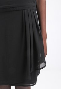 Robe noire avec un design plissé et une superposition transparente au niveau de l'ourlet. Texture de tissu lisse avec une taille cintrée et une silhouette fluide.