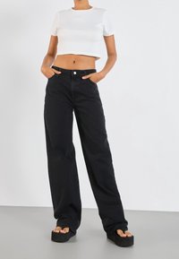 Person med svarta vida jeanstyper, vit kortärmad t-shirt och svarta platåsandaler, står med händerna i fickorna på en grå golv.