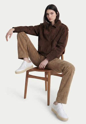 Jeune femme aux cheveux foncés portant une veste et un pantalon en daim marron, des baskets blanches, assise sur une chaise en bois avec une jambe relevée.