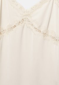 Tissu couleur crème avec bordure en dentelle festonnée formant un décolleté en V et un détail diagonal sur le devant.
