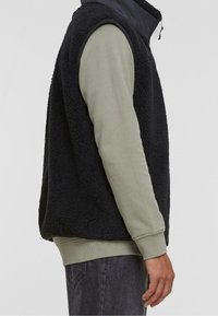 Černá fleecová vesta s vysokým límcem, s měkkou texturou a zapínáním na zip vpředu, nošená přes dlouhý olivově zelený svetr.