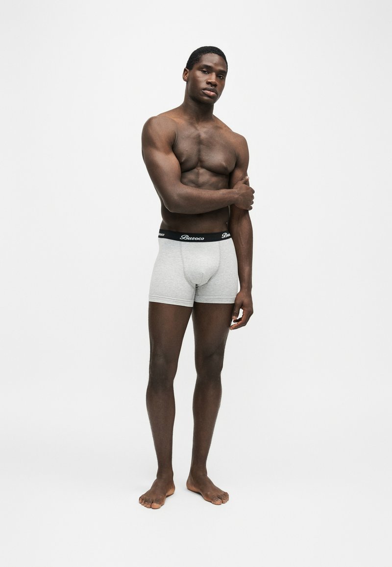 Homme musclé debout pieds nus portant un boxer gris clair avec une ceinture noire, posant avec un bras croisé sur sa poitrine.
