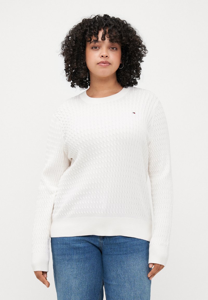 Hvit strikket genser med et teksturert mønster, rund halsuav, og ribbestrikkede mansjetter, kombinert med blå denimjeans.