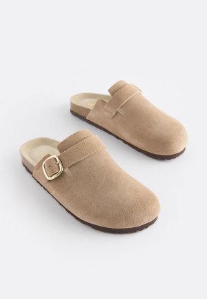 Beige suède klompen met een afgeronde neus, verstelbare riem en goudkleurige gesp. Gestructureerde buitenzool voor grip.