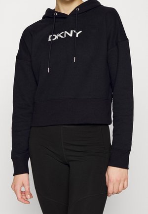 Person trägt einen schwarzen, kurz geschnittenen Hoodie mit "DKNY"-Logo auf der Brust und schwarze Leggings, steht vor einem einfachen hellen Hintergrund.