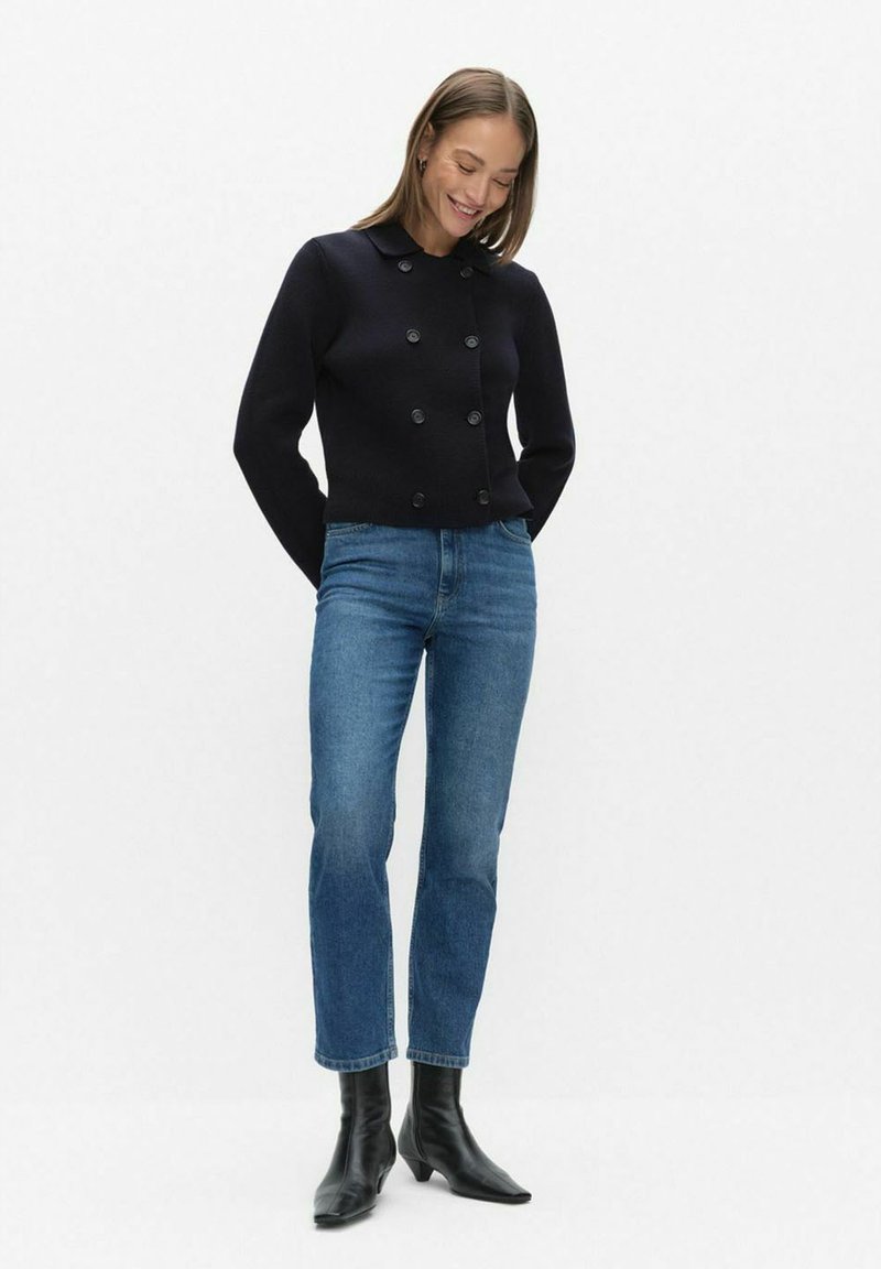 Marineblauwe dubbelgeknoopte gebreide jas met een kraag en grote knopen, gecombineerd met lichtblauwe jeans met rechte pijpen en zwarte enkellaarzen.