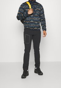 Námorníctví vzorovaný fleecový pulovr s polovičním zipem, spárovaný s černými strečovými kalhotami s bočním kapsou. Černé robustní boty a batoh s žlutými akcenty.