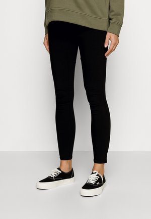 Schwarze Leggings mit glatter Textur und tailliertem Design, kombiniert mit schwarzen Stoffschuhen mit weißen Sohlen und Schnürsenkeln.