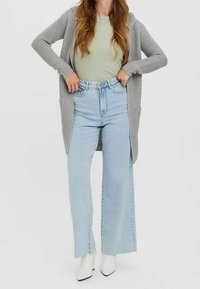 Šedý pletený kardigan přes světlé zelené žebrované tričko, doplněný o světle modré široké džíny a bílé kotníkové boty na podpatku.