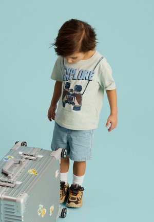 Young child wearing "Explore" shirt and shorts stands looking down at silver suitcase with travel stickers on turquoise background.